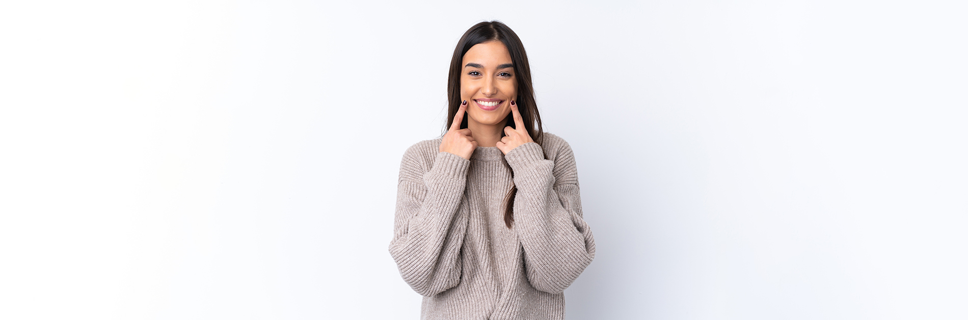 The image is a photograph of a woman with light skin, smiling at the camera. She appears to be in her late twenties or early thirties and has long hair. Her eyes are looking directly at the camera, and she is holding up her index finger near her mouth as if she s making a point or emphasizing something. The background is plain and light-colored, which suggests that this could be a stock photo used for various purposes such as advertising, personal branding, or lifestyle content.