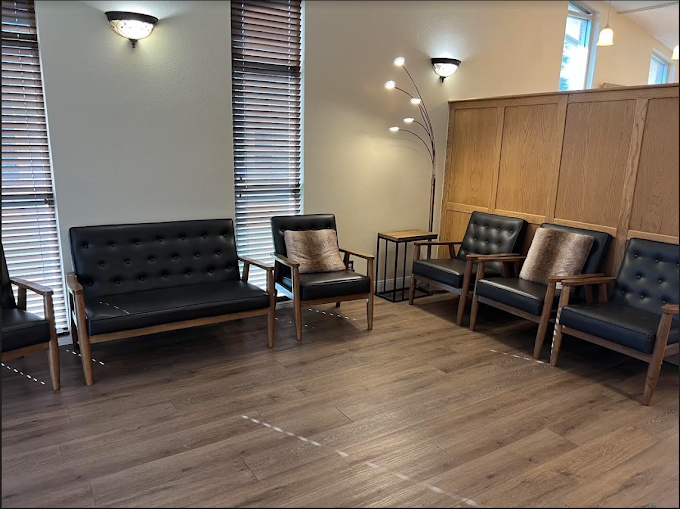 This is an interior photograph showing a waiting area with multiple chairs arranged around a central table, set against a wall-mounted light fixture and a wooden panel. The flooring appears to be a combination of tiles and wood, and there s a large window on the right side of the image allowing natural light into the room.