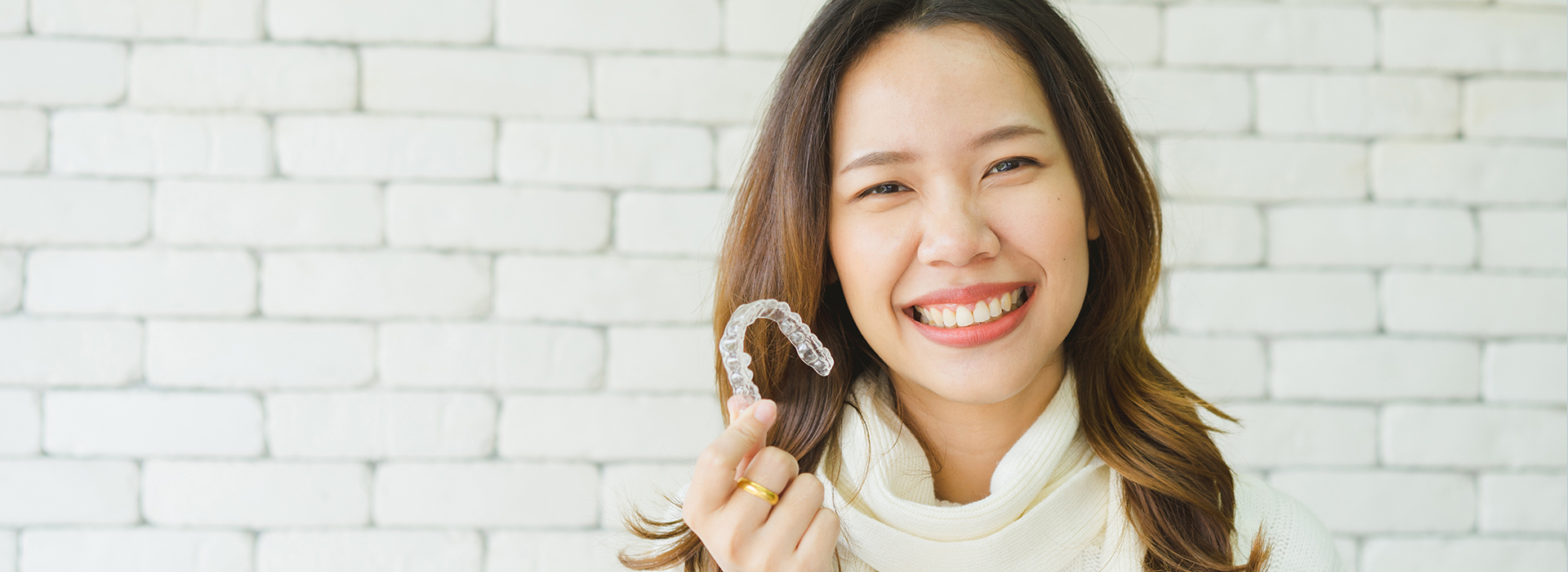 A woman with a smile holds up a toothbrush while standing against a brick wall, appearing happy and engaged.