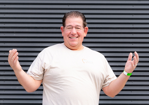 The image shows a man standing with his hands open and palms facing upwards, wearing a casual t-shirt and glasses, against a background of black metal fencing.