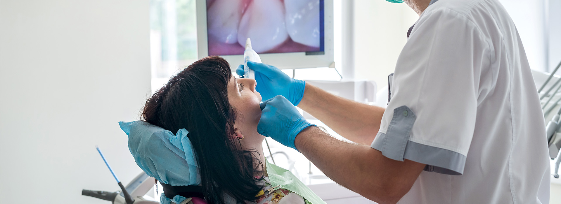 A dental hygienist performing a cleaning procedure on a patient s teeth while wearing protective gloves and a surgical mask, with a dental chair and equipment visible in the background.